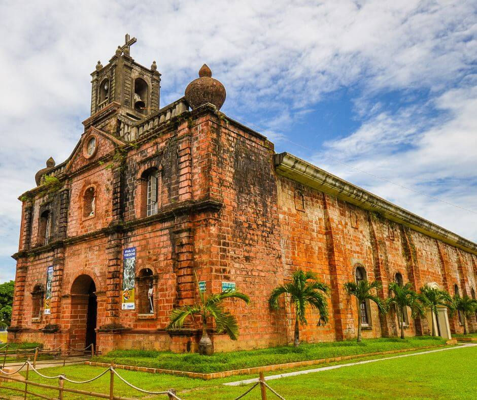 St. Michel Church a historic red-brick church with a cross-topped bell tower under a blue sky. Palm trees line the foreground; banners hang on the facade.