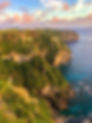 Maùyun Cliff Itbayat a cliffside view of lush, green cliffs and blue ocean under a pink-orange sunset sky. A winding path is visible on the cliffs. Serene mood.