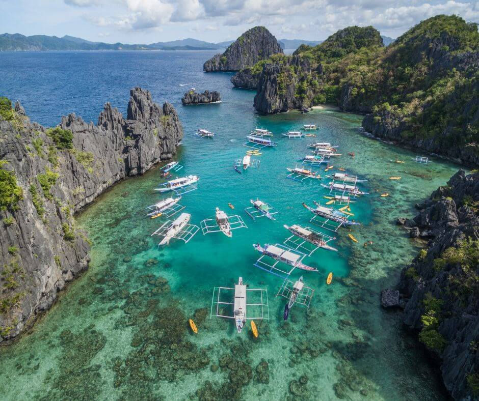 Many outrigger boats in a turquoise lagoon beside steep rocky islands and cliffs under a cloudy sky, calm tropical seascape