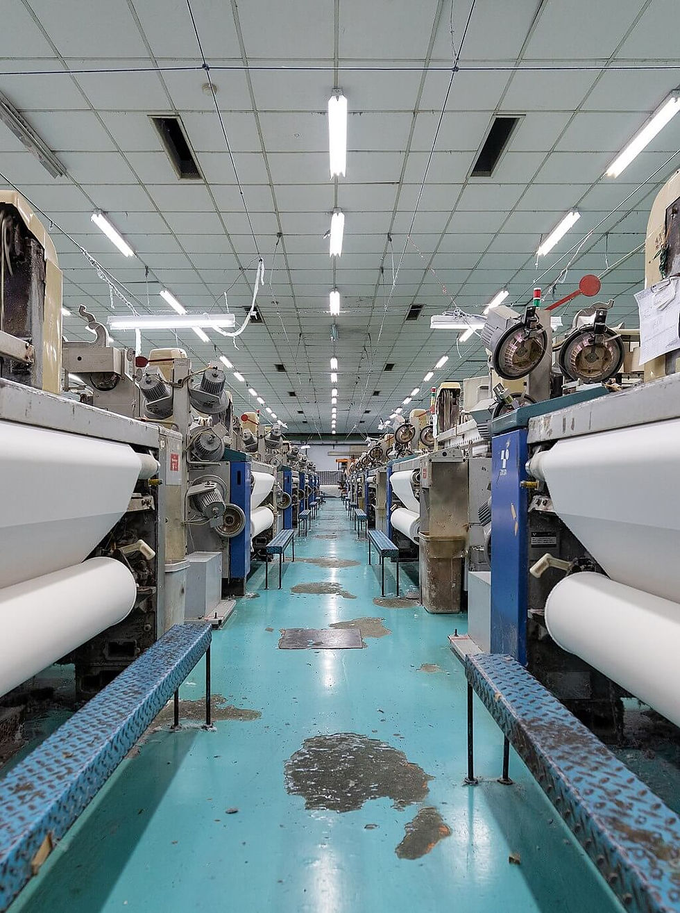 Industrial factory interior with rows of Abaca textile machines on a blue floor, white ceiling lights above, and large rolls of white fabric.