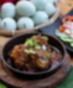 Chicken in brown sauce with green onions in a brown bowl. Background: blue eggs in a basket, sliced cucumbers, and tomatoes. Rustic setting.