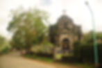 Weathered stone church with a large cross on top, surrounded by lush greenery. A winding road leads past it. Overcast sky sets a serene mood.