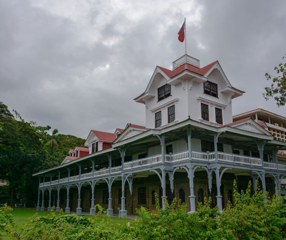 A white colonial-style building with a red roof and a flag on top under cloudy skies, surrounded by greenery.