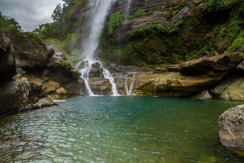 Bomod-Ok Falls a serene waterfall cascade into a clear turquoise pool, surrounded by lush greenery and rocky cliffs under a cloudy sky.