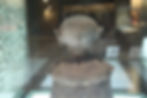 Ancient clay sculpture with a round head and pointed ears displayed in a dimly lit museum, encased in glass. Neutral tones dominate.