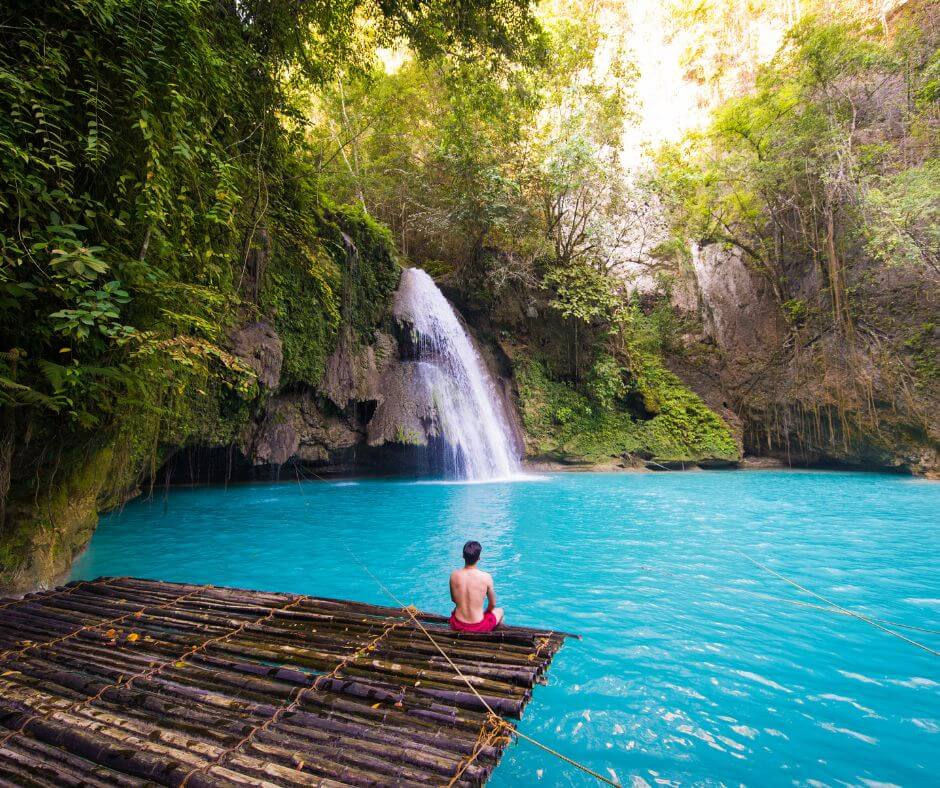 Kawasan Falls in Badian, a person in red shorts sits on a bamboo raft, overlooking a turquoise waterfall in a lush, green forest, creating a serene mood.