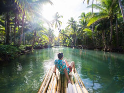 Man sits on a bamboo raft in a calm tropical river, surrounded by lush palm trees and bright sunlight.