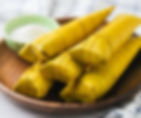 Suman Malagkit , four yellow banana leaf-wrapped parcels on a wooden plate, with a small green bowl of white powder nearby, set on a light surface.
