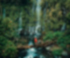 Person in red dress crossing a log bridge in lush green forest with cascading Asik-Asik Waterfall in background, creating a serene, adventurous mood.