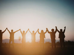 Silhouettes of six people holding hands at sunset, standing in a field with mountains in the background, creating a sense of unity.