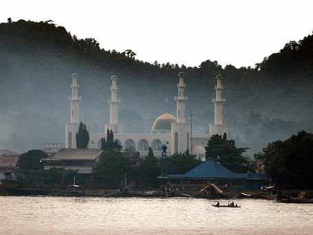 Tulay Mosque with white minarets and a golden dome amidst misty trees, located by water. Boats are visible in the foreground, creating a serene scene.