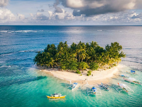 Guyam island in Siargao, a small tropical island with palm trees, surrounded by turquoise water. Several colorful boats docked at the sandy shore under a partly cloudy sky.