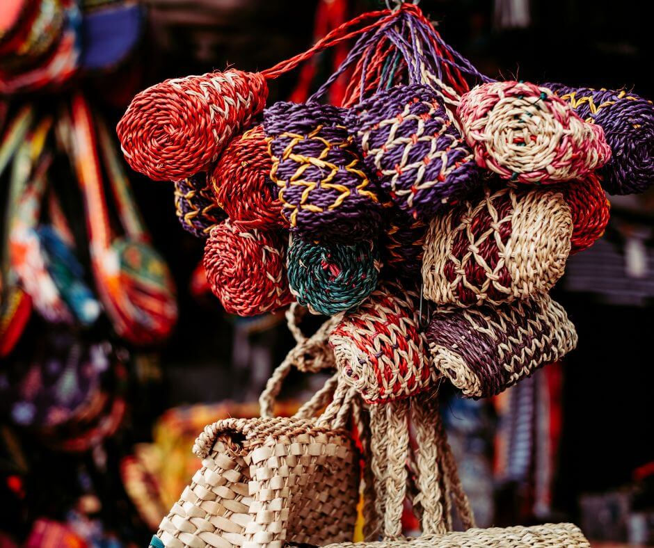 Colorful woven bags hang in a market stall. The bags feature red, purple, and natural hues with intricate patterns, evoking a vibrant mood.