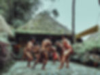 Men in traditional attire dance with gongs in a village setting. Thatched huts and lush greenery surround them. Sign reads: "TAM•AWAN VILLAGE".