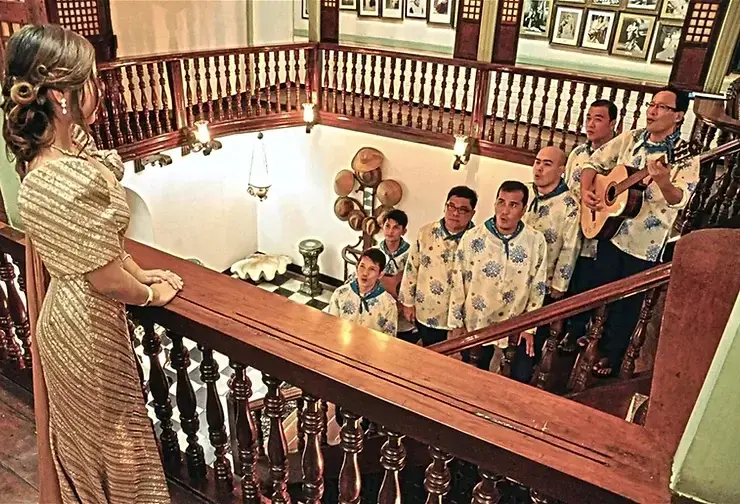 Harana, a woman in gold attire looks over a wooden balcony at a group of men in patterned shirts, one playing guitar, in a warmly lit vintage setting.
