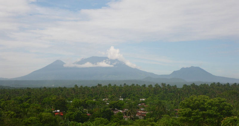 Green landscape with houses and palm trees in the foreground. A distant mountain range with clouds covers one peak. Sky is partly cloudy.