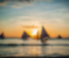 Sailboats with blue sails glide across calm sea at sunset, golden light reflecting on the water under a partly cloudy sky.
