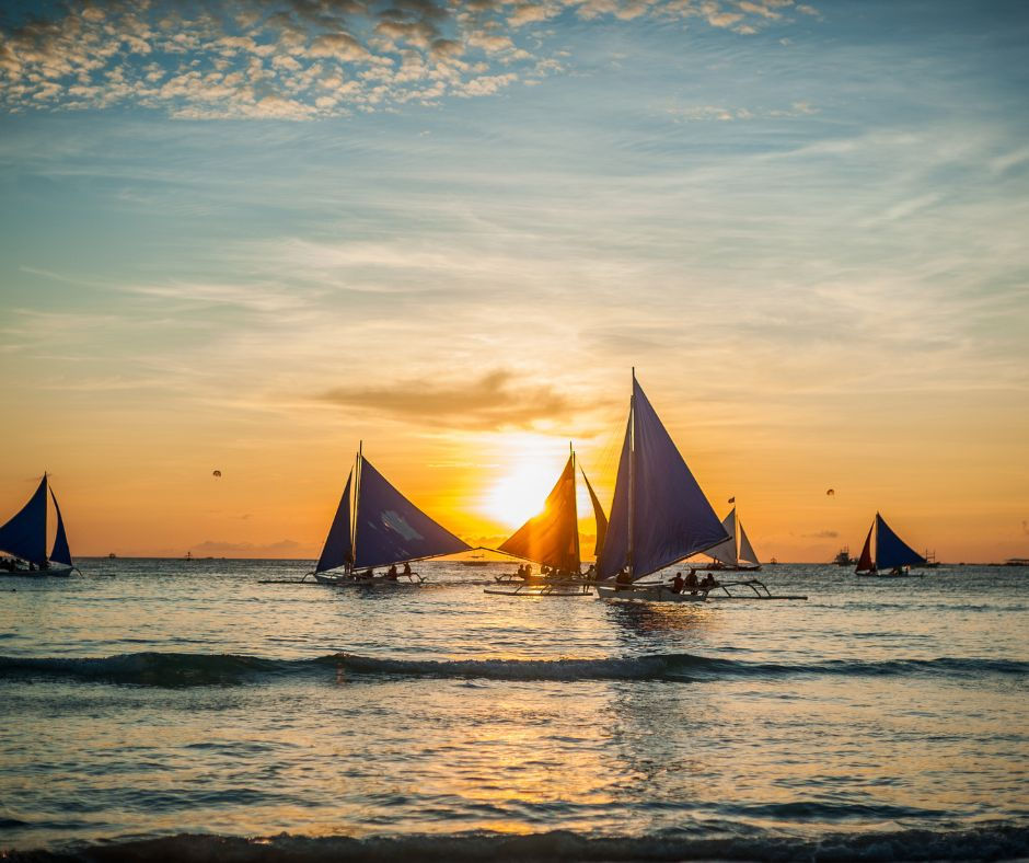 Sailboats with blue sails on calm sea at sunset, silhouetted against an orange sky with golden reflections.