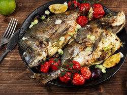 Grilled fish on a black plate garnished with roasted tomatoes, lemons, and scallions. Wooden table setting with a lime and cutlery.