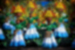 Festive dancers in green tops and layered blue skirts, holding colorful fruit arrangements, perform outdoors with joyful expressions.