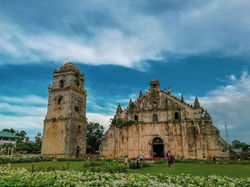 Historic stone church and bell tower with tourists on a lush, flower-filled lawn. Blue sky overhead creates a serene mood.