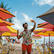 Man wearing floral shirt and orange shorts taking a selfie on a lively beach. Colorful umbrellas and chairs line the sandy shore under blue sky.