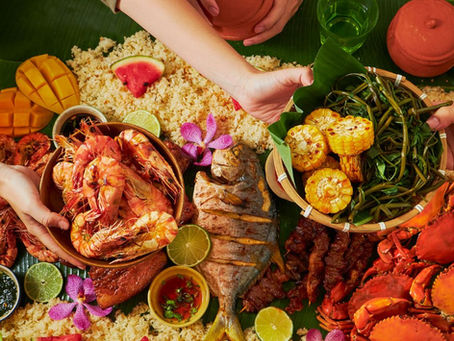 A festive spread with seafood, grilled fish, corn, and vegetables on banana leaves. Hands serving, vibrant colors, and decorative flowers.