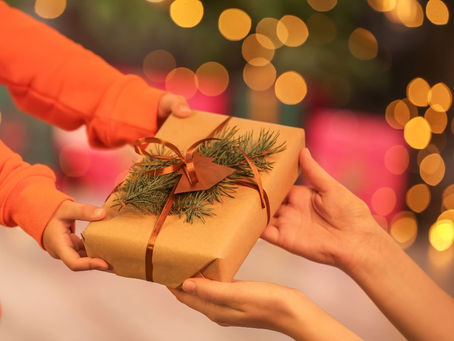 Hands exchange a gift wrapped in brown paper with pine and ribbon accents. Background has warm, festive lights and soft bokeh effect.