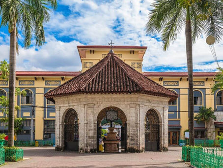 Historic stone structure with a red-tiled roof and arches in a plaza. Palm trees and a yellow building in the background under a blue sky.