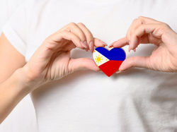 Hands gently hold a heart-shaped pin with the Philippine flag. The person wears a white shirt, set against a plain background.
