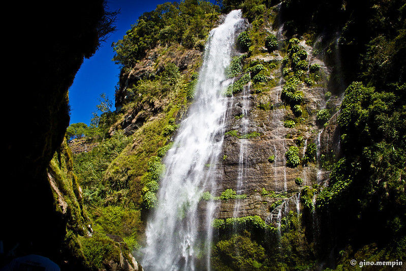 Bomod-ok Falls a tall waterfall cascading over a lush, green cliff under a clear blue sky. Vegetation surrounds the area, creating a scenic and serene mood.