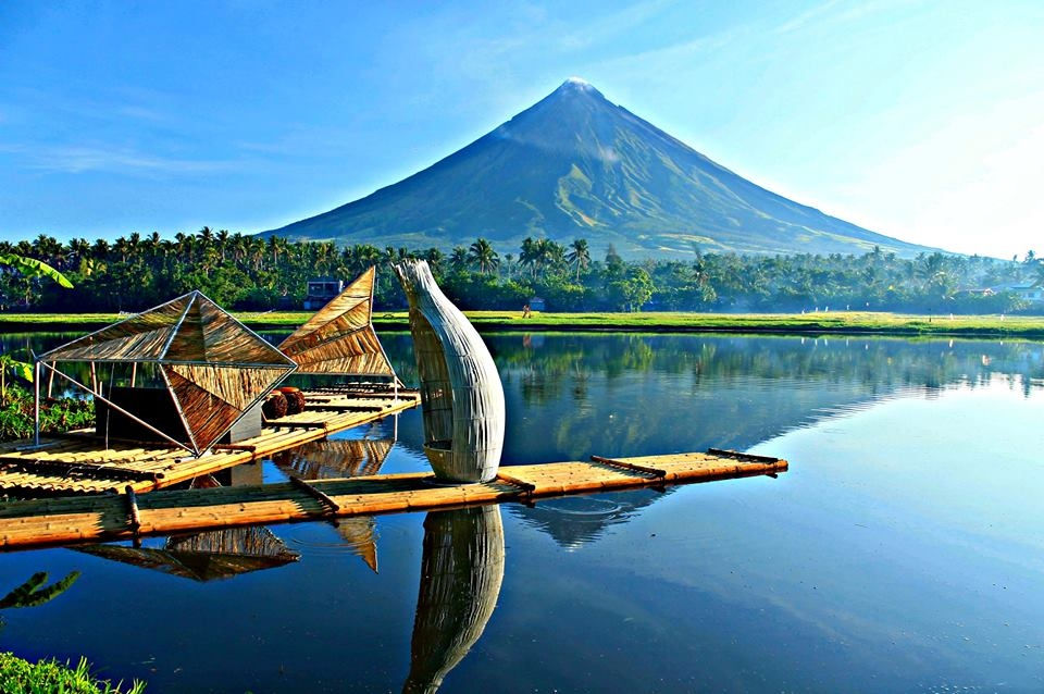 Bamboo structures float on a calm lake with a reflected view of Mayon volcano in the background. Bright, clear sky and lush greenery.