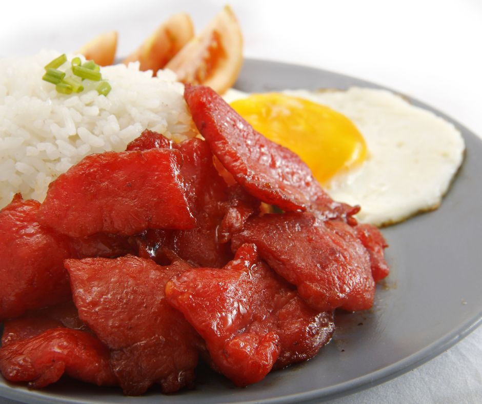 Plate of red glazed meat, fried egg, rice with chives, and tomato slices on gray plate. Bright and appetizing breakfast setting.