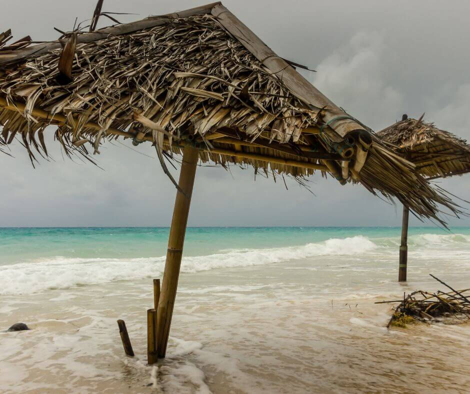 Waves crash on a beach with tilted thatched umbrellas under a gray sky. A moody, stormy atmosphere, turquoise sea in the background.