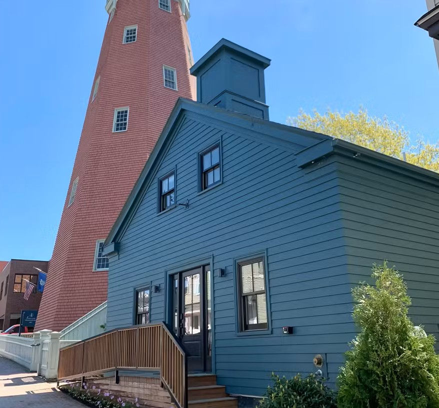Classic exterior of Best Bower guesthouse at the peak of Munjoy Hill beside the iconic red Portland Observatory, with blue siding, entry stairs, and clear blue sky in Portland, Maine.