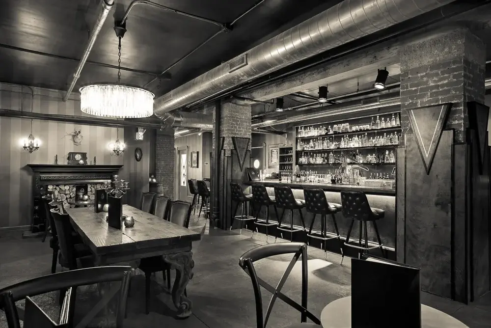 Wide interior perspective of The Velveteen Speakeasy showing a long wooden table, tufted seating, a decorative fireplace mantel, and the bar in the background under exposed ductwork and industrial-chic lighting.