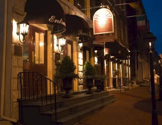 Cozy nighttime view of the main entrance to Penn’s View Hotel along a brick sidewalk in Philadelphia, with lit lanterns, black awnings, potted plants, and glowing signage for the hotel and Panorama restaurant.