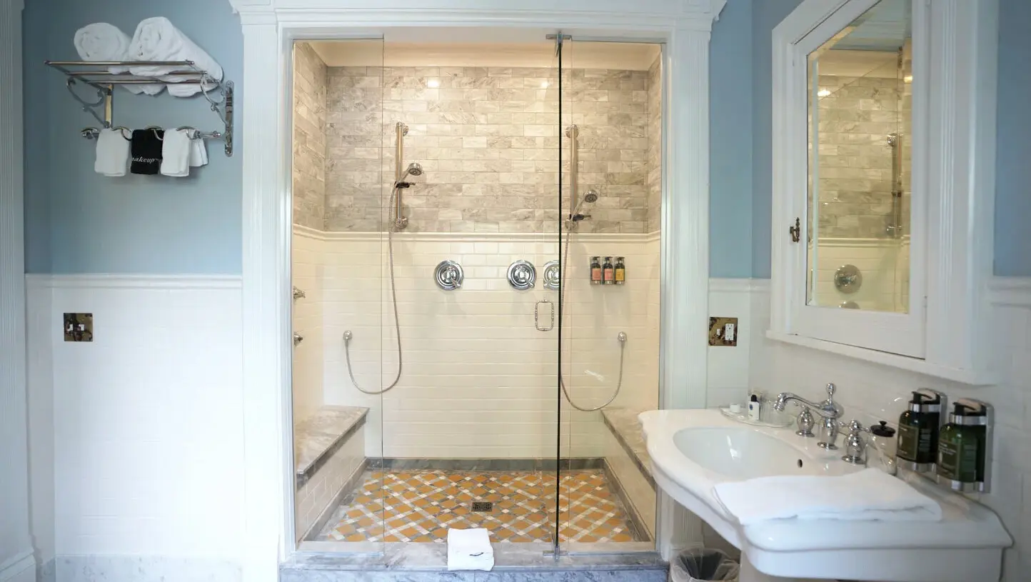 Elegant and spa-like private bathroom interior at Ann Bean Mansion Bed & Breakfast featuring a large walk-in glass shower enclosure with multiple chrome shower heads, marble-tiled walls, and a colorful patterned mosaic tile floor, paired with a classic white pedestal sink with chrome faucet on the right, a wall-mounted mirror cabinet, rolled white towels on a chrome rack, light blue upper walls with white wainscoting and marble trim, and bright, clean lighting throughout the refined space.