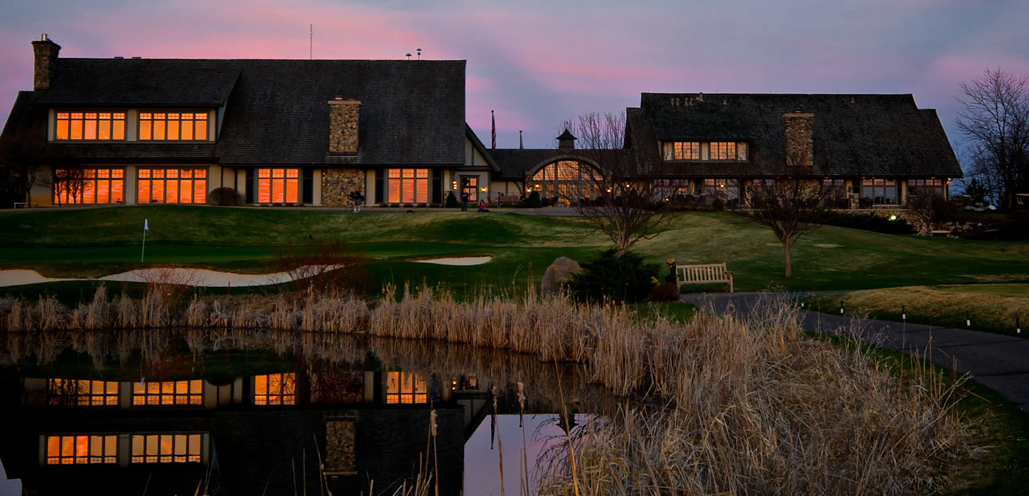 Dramatic twilight view of the elegant stone-and-wood clubhouse at Rush Creek Golf Club glowing with warm interior lights, perfectly reflected in a calm pond with pink and purple sunset skies, highlighting the pristine grounds and upscale ambiance.