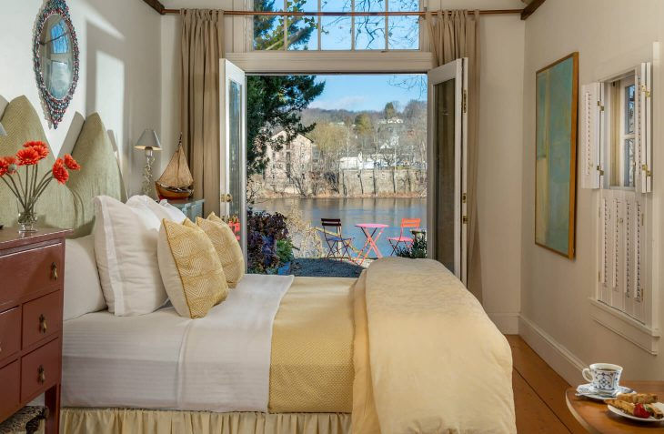 Bright and inviting luxury guest bedroom featuring a large bed dressed in crisp white linens with a pale yellow throw and patterned pillows, a warm wooden dresser topped with a vase of vibrant orange flowers, and open French doors revealing a private balcony with pink chairs overlooking the scenic Delaware River, trees, and distant buildings under a clear blue sky; a small table in the foreground holds a teacup and breakfast plate.