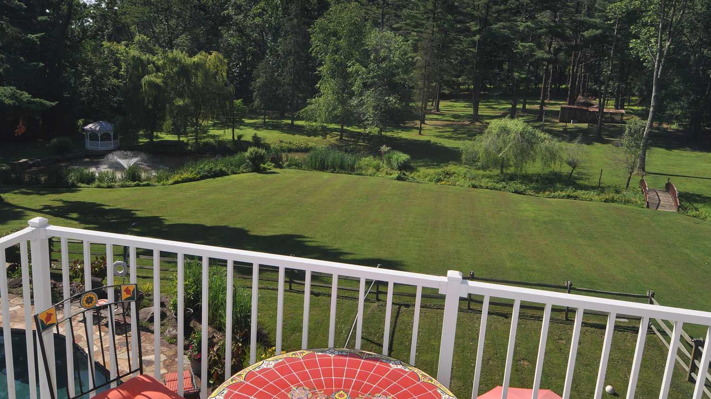 Elevated daytime view from a white-railed balcony at The Inn at Bowman’s Hill overlooking the expansive 5-acre grounds, featuring a vast manicured green lawn, pond with fountain, white gazebo, lush gardens, mature trees and forest backdrop, wooden bridge, with a colorful red tiled table in the foreground and the swimming pool area visible below.