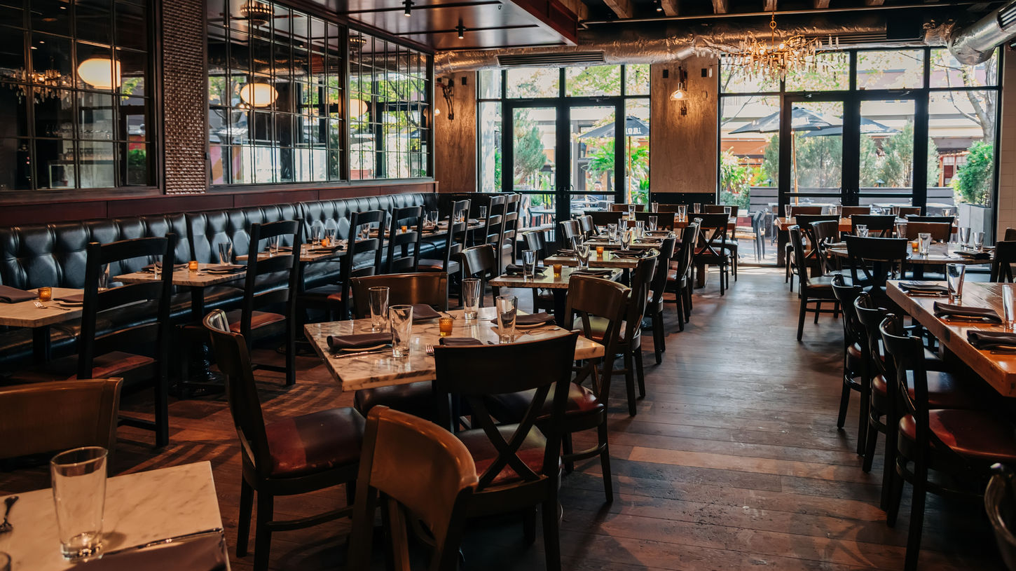 Wide interior view of the main dining room at The Butcher’s Tale showing wooden tables, dark leather booths, marble tabletops, large windows, and exposed ceiling beams in a warm, sophisticated atmosphere.