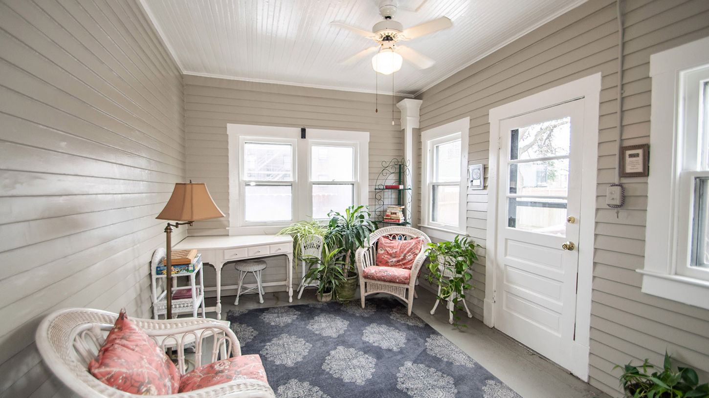 Inviting enclosed sunroom porch at Looking Glass Inn featuring white wicker chairs and loveseat with vibrant red patterned cushions, a white desk/console table, numerous potted plants, a dark patterned area rug on the floor, white beadboard walls and ceiling, a ceiling fan with light fixture, large windows letting in natural light, and a white door leading outside.