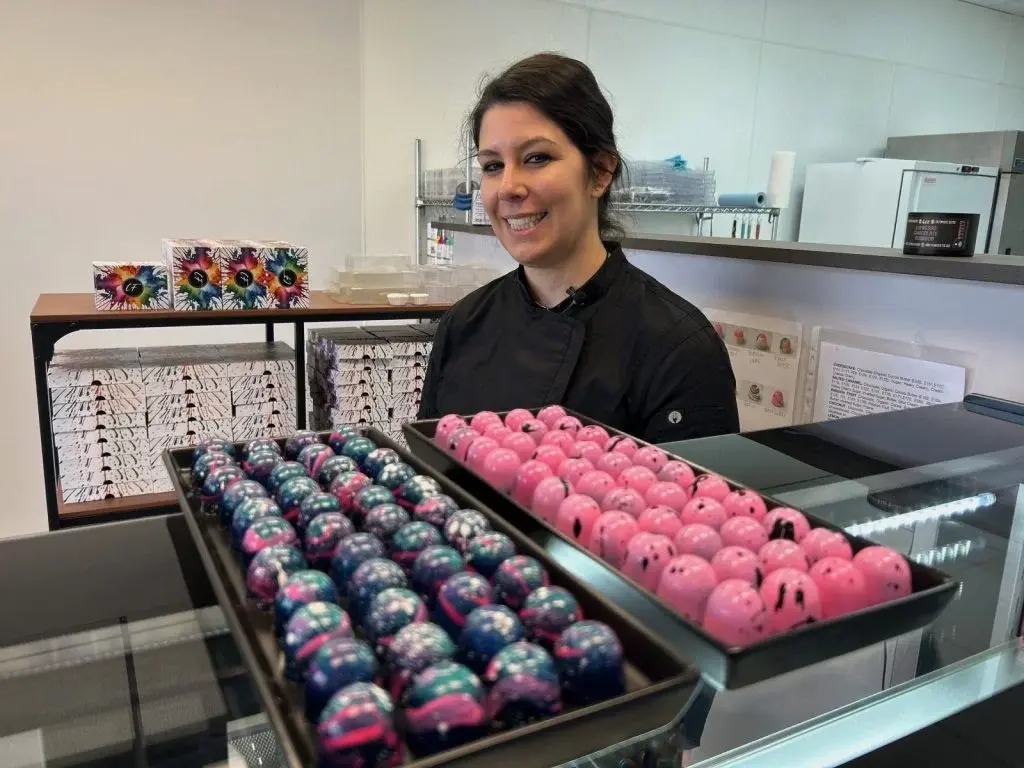 Smiling chocolatier in black chef coat standing behind glass display cases filled with trays of vibrant pink, blue, and multicolored hand-painted bonbons inside the clean, well-lit Cocoa Flake shop in Robbinsdale.