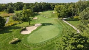 Stunning high-resolution aerial photograph of a classic Pete Dye-designed hole at The Fort Golf Course in Indianapolis, showcasing a beautifully manicured undulating green guarded by multiple large, sculpted sand bunkers, a generous rolling fairway approach, winding cart path, and lush mature trees framing the hole within the historic Fort Harrison State Park on a bright sunny day.