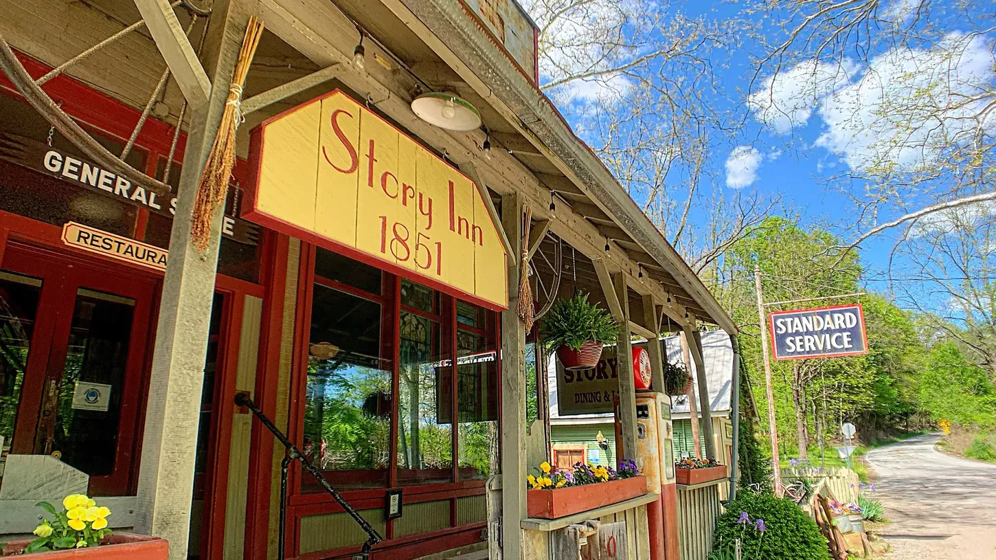Charming weathered exterior of the Story Inn 1851 building with red doors, vintage signage, flower boxes, and the iconic “Story Inn” sign under a bright blue sky with trees in the background.