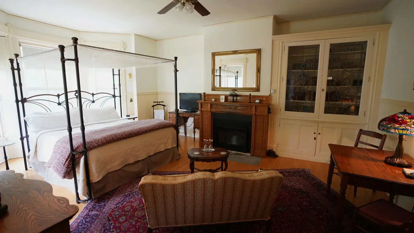 Spacious and sunlit guest suite interior at Ann Bean Mansion Bed & Breakfast featuring a dramatic black wrought-iron four-poster canopy bed with white netting and patterned bedding on the left, a large ornate red-and-blue Persian-style rug covering the hardwood floors, a striped upholstered sofa in the foreground, a wooden fireplace mantel with a large gold-framed mirror above it, a tall built-in glass-door cabinet on the right, a wooden desk with a colorful Tiffany-style stained-glass lamp, and a ceiling fan overhead.