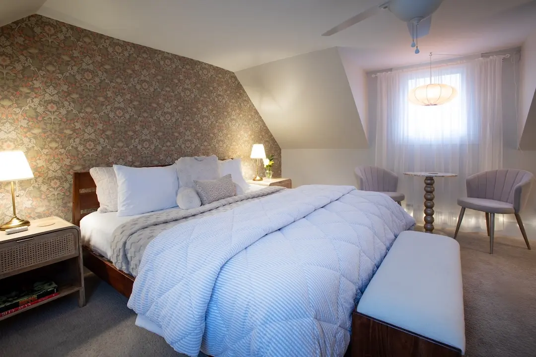 Bright and inviting guest suite at Prairie Guest House featuring a large bed dressed in crisp white linens with a soft blue-and-white striped quilt, multiple plush pillows, and a wooden headboard set against a dramatic floral wallpaper accent wall; a white upholstered bench sits at the foot of the bed, while two gray upholstered chairs and a small round table are positioned near large windows with sheer curtains, all under a sloped ceiling with a ceiling fan and pendant light.