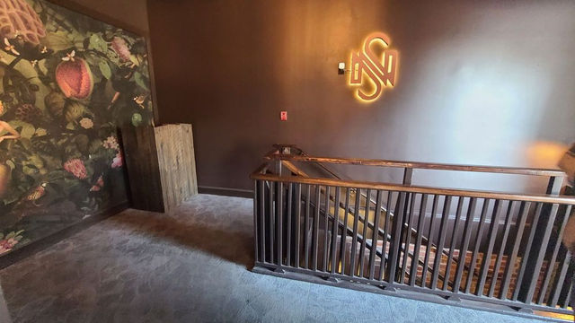 Elegant entrance and staircase at Nowhere Special Cocktail Lounge featuring a large, vibrant floral and fruit mural on the left wall, glowing neon “NS” logo on the dark wall, wooden accents, metal railing, and steps leading down into the sophisticated basement speakeasy lounge.  