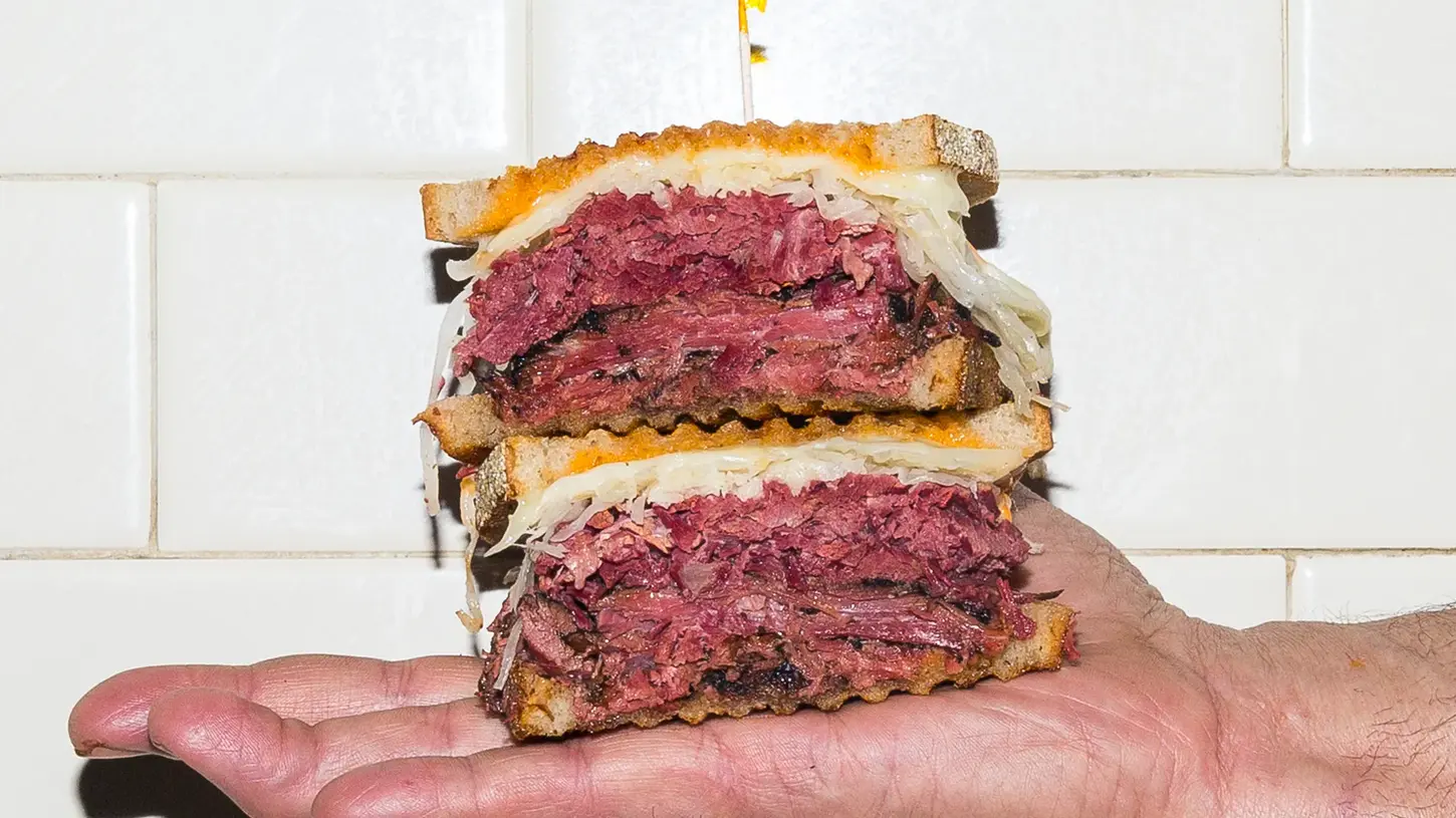 Stacked pastrami sandwich on rye bread held in hand, overflowing with thick layers of house-cured pastrami, sauerkraut, and melted cheese against white tiled background.